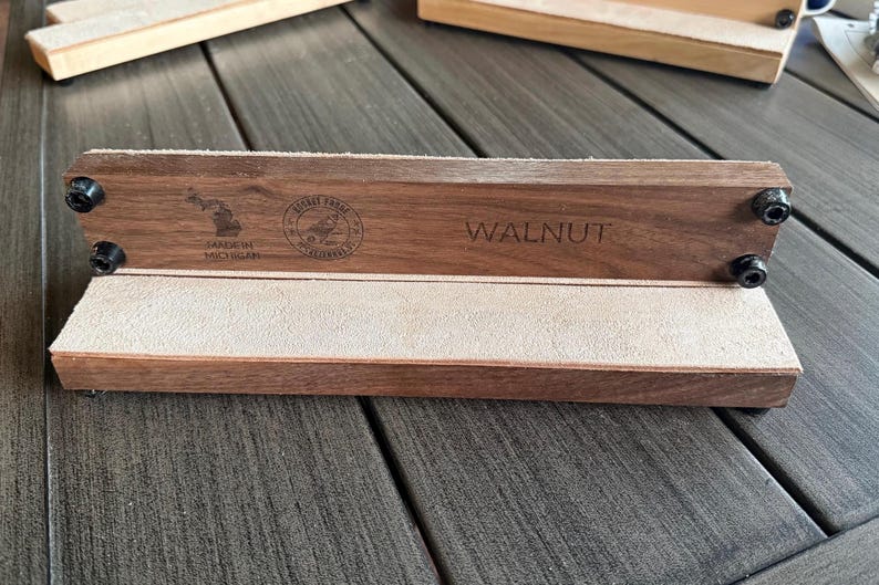 May include: A walnut wood desk organizer with a light-colored suede interior. The organizer has the word "WALNUT" engraved on the front. The text "Made in Michigan" and a logo are also visible. Black screws secure the wood.