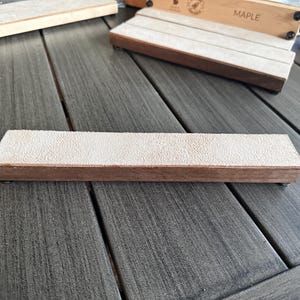 May include: A wooden sharpening block with a light tan abrasive surface. The block is rectangular with a brown wooden base. Other similar blocks are visible in the background. The word "MAPLE" is printed on a wooden box.