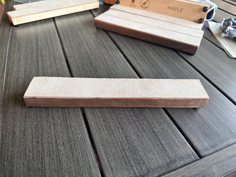 May include: A wooden sharpening block with a light tan abrasive surface. The block is rectangular with a brown wooden base. Other similar blocks are visible in the background. The word "MAPLE" is printed on a wooden box.
