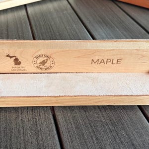 May include: A light-colored maple wood strop with a light-colored suede surface. The wood is engraved with the word "MAPLE", a Michigan state outline, and a rocket logo. The strop is held together with black hardware.