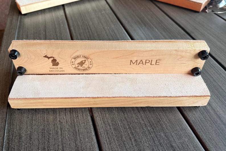 May include: A light-colored maple wood strop with a light-colored suede surface. The wood is engraved with the word "MAPLE", a Michigan state outline, and a rocket logo. The strop is held together with black hardware.