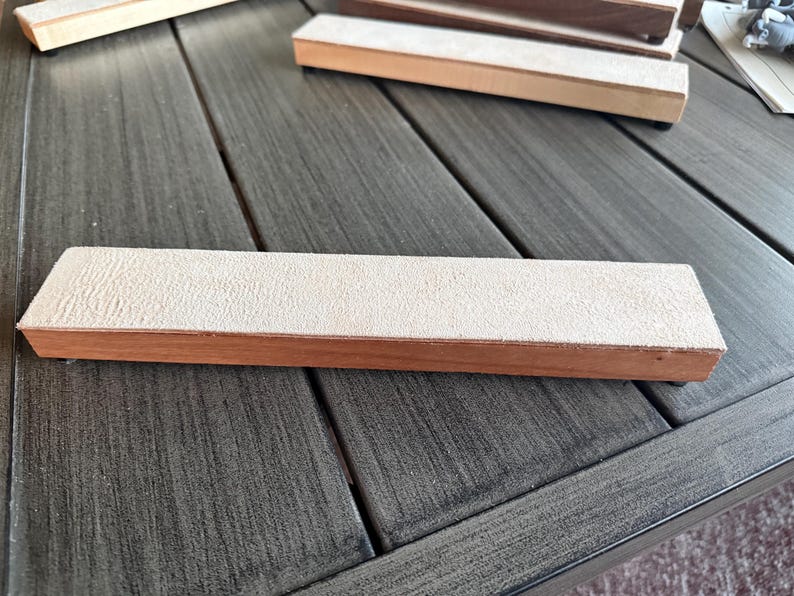 May include: A rectangular sharpening stone with a light tan, textured surface and a wooden base. Several similar stones are visible, suggesting a set or collection of woodworking tools.