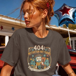 May include: A dark grey t-shirt featuring a graphic print of a tired computer monitor character with messy hair, surrounded by coffee mugs and the text 404: MOTIVATION NOT FOUND, EST. 2026 | SINCERELY, A DEFLATED GENIUS, and large 404 numbers.
