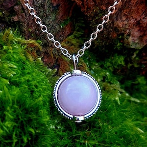 Op de afbeelding: Een zilveren ketting met een ronde, lichtroze hanger. De hanger is gezet in een zilveren frame met een touwachtige rand. De ketting wordt getoond op een met mos bedekte stam, met een vage achtergrond van bladeren.