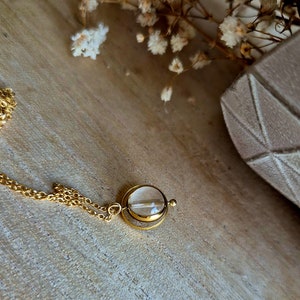 May include: A gold-colored necklace with a clear, circular pendant. The pendant has a small, clear stone inside. The necklace is on a wooden surface, with dried flowers in the background. The necklace is a piece of jewelry.