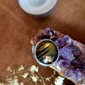 Könnte beinhalten: Ein silberner Ring mit einem großen, runden, dunklen Edelstein mit gelben und schwarzen Streifen. Der Ring ist auf einer lila Kristallformation platziert, mit einer weißen Kerze und getrockneten Blumen im Hintergrund. Der Ring ist ein Schmuckstück.