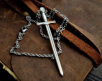 May include: A silver sword pendant necklace with a detailed blade and hilt, set on a brown leather surface. The chain is also silver, and the pendant has a small, clear gem at the top. The background includes a geode and a rolled leather strap.