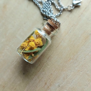 Könnte beinhalten: Ein kleiner Anhänger aus einer klaren Glasflasche an einer silberfarbenen Metallkette. Die Flasche ist mit einem Korken verschlossen und enthält konservierte gelbe Mimosenblüten sowie grüne farnartige Blätter als botanisches Arrangement.