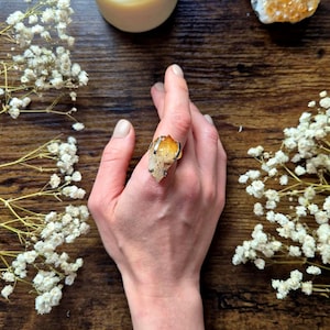 Könnte beinhalten: Eine Hand mit einem silbernen Ring mit einem großen, rohen, orangefarbenen Citrin-Kristall. Der Ring befindet sich auf einer dunklen Holzoberfläche, mit einer brennenden Kerze und getrockneten weißen Blumen. Ein Stück Citrin ist ebenfalls sichtbar.