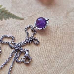 Può includere: Una collana d'argento con un pendente rotondo di ametista viola scuro. È presente anche una pietra più piccola, rosa chiaro. La catena è collegata e ha una chiusura. Lo sfondo è una superficie strutturata marrone chiaro.