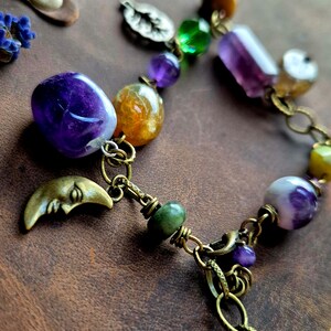 May include: A handcrafted bracelet featuring a variety of colorful gemstones, including amethyst, citrine, and green jade. The bracelet has a bronze chain and a crescent moon charm. The beads are connected by bronze links.