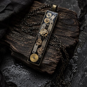 Può includere: Un ciondolo rettangolare caratterizzato da un assortimento di ingranaggi di orologi e componenti meccanici in metallo color oro e argento incastonati in una base di resina scura, appeso a una catena a maglie in metallo color bronzo.