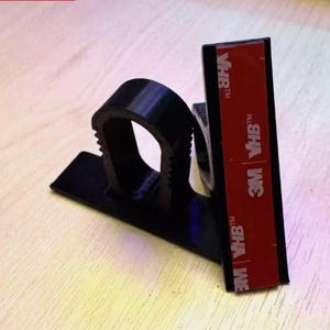 May include: A black plastic cable clip with a curved opening and a flat base. A red and white 3M VHB adhesive strip is attached to the back of the clip, ready for mounting. The clip is designed to organize and secure cables.