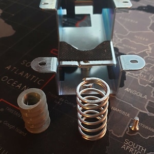May include: A close-up of a metal spring, a metal bracket, and a metal cylinder. The metal bracket is attached to a black surface. The metal cylinder is made of multiple stacked rings. The metal spring is coiled and has a silver finish.