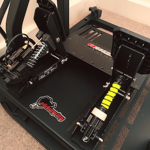 Könnte beinhalten: Schwarzes Rennsimulator-Pedalset mit Nürburgring-Logo. Die Pedale haben Federn und gelb-schwarze Stoßdämpfer. Das Set ist auf einem schwarzen Metallrahmen montiert.