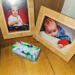 Può includere: Due cornici per foto in legno con facce in vetro, ciascuna delle quali mostra una foto di un bambino piccolo. La cornice a sinistra mostra un ragazzo con le cuffie e una camicia bianca. La cornice a destra mostra un bambino che indossa una camicia a quadri rossi e neri. Una saponetta rettangolare colorata si trova in primo piano.