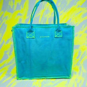 May include: A teal tote bag with a rectangular shape and two handles. The bag has neon green stitching and accents. The text "EEVHUN" is printed on the front of the bag. The background is a mix of gray and yellow.