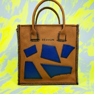 Peut inclure: Sac fourre-tout marron avec deux anses et le texte "EEVHUN" imprimé sur le devant. Le sac présente des patchs géométriques bleus cousus sur le devant. Le sac a une fermeture éclair et une bordure cousue noire.
