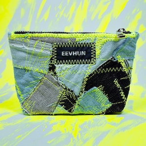 Può includere: Piccola pochette in patchwork di denim con chiusura a zip. È realizzata con ritagli di denim blu e nero, cuciti insieme con filo giallo neon. Un'etichetta nera con la scritta "EEVHUN" è cucita sul davanti.