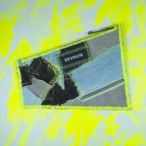 Può includere: Una piccola pochette rettangolare in denim con una cerniera e un bordo di colore giallo fluo. La pochette è realizzata con diversi pezzi di denim cuciti insieme in un design patchwork. Una piccola etichetta nera con il testo "EEVHUN" è cucita sulla parte anteriore della pochette.
