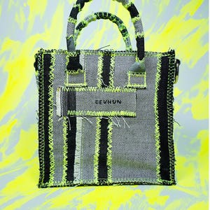 Può includere: Una borsa tote in denim grigio con strisce verticali nere e cuciture verde neon. La borsa ha due manici e una piccola etichetta con la scritta "EEVUNN". Lo sfondo è un mix di giallo e grigio.