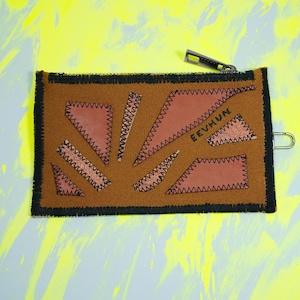 Può includere: Un astuccio rettangolare marrone con bordo nero e chiusura a cerniera. L'astuccio presenta diverse toppe marroni chiare di forma irregolare cucite sulla superficie con filo nero. La parola "EEVHUN" è cucita su una delle toppe.