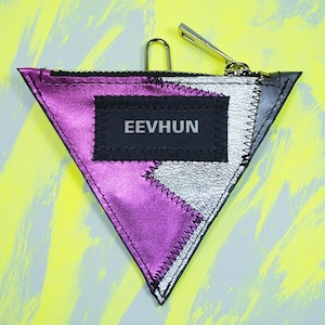 Può includere: Un portamonete triangolare con una combinazione di colori viola metallizzato, argento e nero. Il portamonete ha una chiusura a zip e un piccolo fermaglio in metallo. La parola "EEVHUN" è stampata su una toppa rettangolare nera.