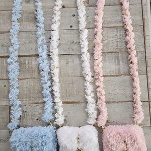 May include: Three small, fuzzy crossbody bags in light blue, white, and pale pink. Each bag features a matching long, textured strap, a gold-toned metal clasp closure on the front flap, and a small gold-toned rectangular logo plate at the bottom.