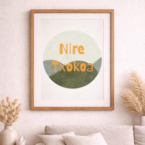 Op de afbeelding: Een ingelijste kunstdruk met de woorden "Nire Txokoa" in oranje letters. De tekst staat tegen een cirkelvormige achtergrond met groene en grijze bergachtige vormen. Het frame is gemaakt van lichtbruin hout.