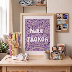 Op de afbeelding: Een ingelijste kunstdruk met de tekst "NIRE TKOKOA" in wit over een paars en lavendel abstract ontwerp. De print staat op een houten bureau met een koptelefoon, boeken en decoratieve items.