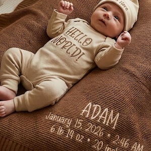 May include: A baby in a beige onesie and hat, with "HELLO WORLD!" and "I'm Here" printed on them, rests on a brown knitted blanket. The blanket is embroidered with the baby's name, birthdate, time, weight (3.0 kg), and length (50.8 cm).