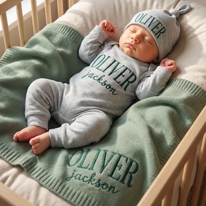 Peut inclure: Un bébé endormi dans un berceau en bois, vêtu d'un body et d'un bonnet gris brodés de "OLIVER" et "jackson" en vert. Le bébé est allongé sur une couverture verte avec la même broderie. Un mobile avec des étoiles et une lune est suspendu au-dessus du berceau.