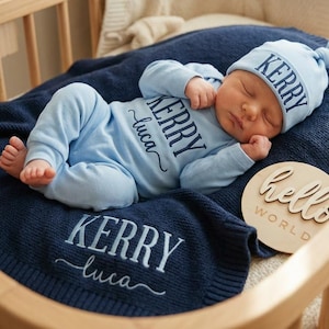 Pode incluir: Um bebê dormindo usando um traje azul claro e chapéu combinando, deitado em um cobertor azul marinho dentro de um berço de madeira. O cobertor e o chapéu são bordados com o nome "Kerry Luca". Uma placa de madeira diz "hello WORLD".
