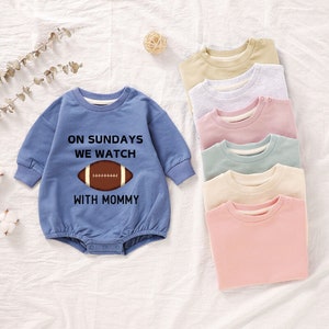 Pode incluir: Um body azul de manga comprida com o texto "On Sundays We Watch With Mommy" e um gráfico de futebol americano marrom. O body é mostrado com outros cinco bodys em cores diferentes.