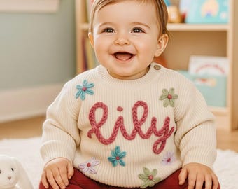 Puede incluir: Un suéter de bebé color crema con el nombre "Lily" bordado en hilo rosa. El suéter está decorado con aplicaciones de flores de colores. El bebé lleva mallas burdeos y está sentado sobre una alfombra blanca.