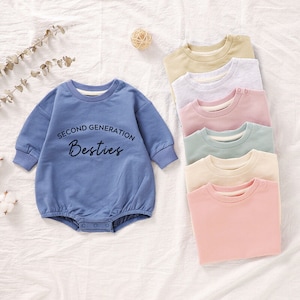 May include: A set of six baby onesies in various colors, including light blue, pink, beige, and gray. One onesie features the text "Second Generation Besties" in black.