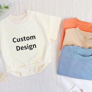 May include: A cream baby romper with the text "Custom Design" in black. Beside it are three short-sleeved tops in orange, beige, and blue. The garments are made of soft material and are suitable for babies and toddlers.