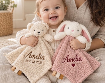 Può includere: Due doudou in peluche, uno con un orsetto color crema e uno con un coniglietto rosa polvere. Entrambe le coperte hanno un tessuto testurizzato tipo sherpa e sono ricamate con il nome Amelia. L'orso riporta la data DEC 12, 2025, e il coniglio un cuore.