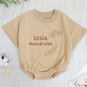 May include: A light brown short-sleeved bodysuit with a graphic of a mandrake plant and the text "little mandrake".