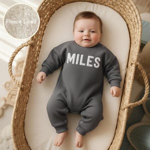 May include: A baby wearing a dark gray fleece-lined jumpsuit with the name "MILES" in white block letters. The baby is lying in a woven wicker bassinet with a cream-colored mattress. The jumpsuit has snap closures at the legs.
