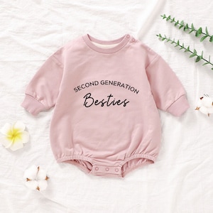 May include: A pink long-sleeved baby onesie with the text "Second Generation Besties" printed on the front.