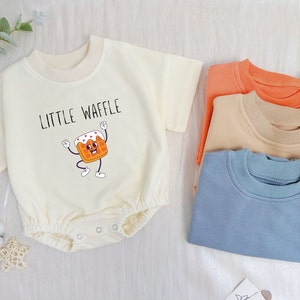 Op de afbeelding: Een witte baby romper met korte mouwen en een afbeelding van een cartoonwafel met de tekst "Little Waffle".