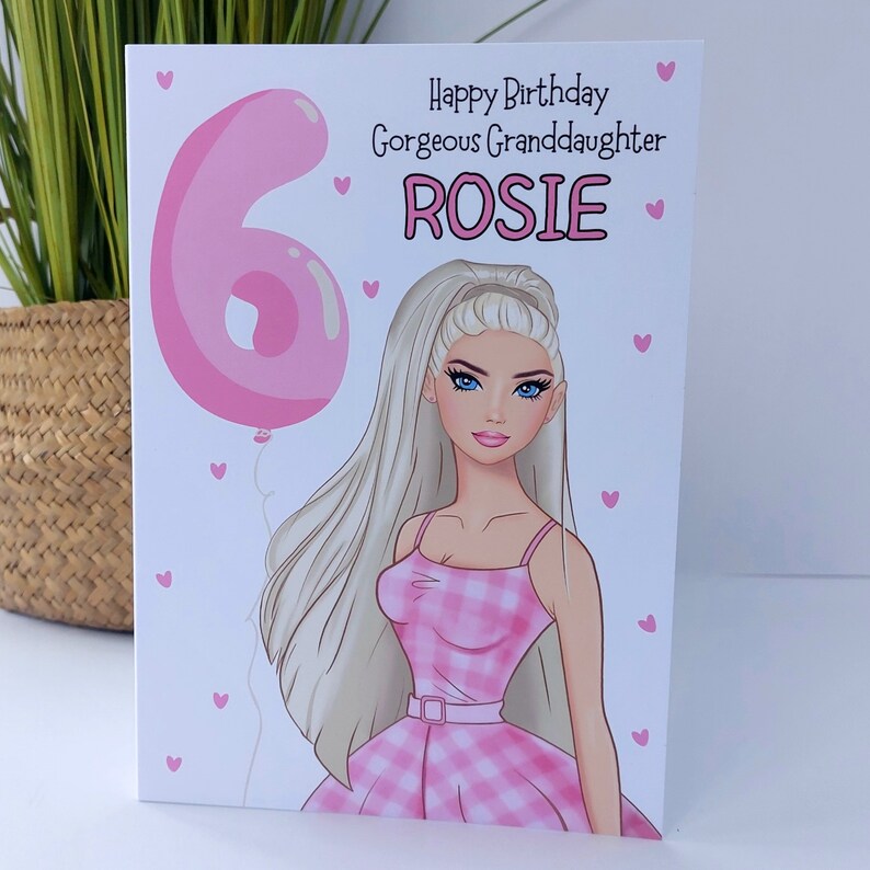 May include: A birthday card featuring a cartoon illustration of a woman with long blonde hair and blue eyes, wearing a pink gingham dress. The card reads "Happy Birthday Gorgeous Granddaughter ROSIE" with a pink number 6 balloon.