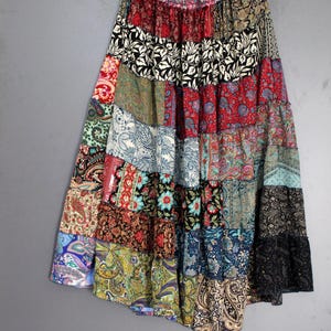 May include: A patchwork skirt with a tiered design, featuring a variety of colorful patterns and fabrics. The skirt hangs from a wooden hanger against a gray background. The skirt's design includes paisley, floral, and geometric prints in shades of red, blue, green, and black.