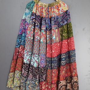 Könnte beinhalten: Ein langer Patchwork-Rock mit Stufen, der eine Vielzahl farbenfroher Paisley- und Blumenmuster aufweist. Der Rock besteht aus mehreren Stoffbahnen in Rot-, Blau-, Grün- und Goldtönen. Er hängt an einem Holzbügel.