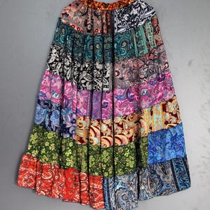Könnte beinhalten: Ein langer Patchwork-Rock mit mehrschichtigem Design, der verschiedene Paisley- und Blumenmuster in leuchtenden Farben aufweist. Der Rock hängt an einem Holzbügel vor grauem Hintergrund. Der Saum des Rocks hat einen gerüschten Rand.