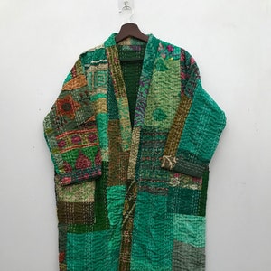 Puede incluir: Una bata tipo kimono de patchwork en tonos verdes, marrones y rosas. La bata está hecha de varias piezas de tela cosidas con hilo blanco. La prenda está colgada de una percha de madera sobre un fondo blanco.