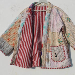 Puede incluir: Una chaqueta acolchada de patchwork con un forro a rayas rojo y blanco. La chaqueta tiene un corte holgado y está hecha de una variedad de telas en tonos beige, rosa y azul.