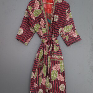 May include: A patterned robe in red, black, and green, with a tie waist. The robe features a leaf and swirl design. It is hanging on a wooden hanger against a grey background. The robe has three-quarter sleeves.