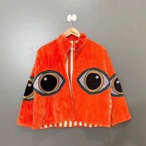 Könnte beinhalten: Orangefarbene Samtjacke mit großen, gestickten Augendesigns auf der Vorderseite und den Ärmeln. Die Jacke hat einen hohen Kragen und ein gestreiftes Futter. Die Augen sind schwarz, gold und lila.